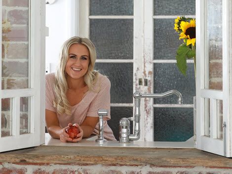 Leisa Tuite leaning on the sill of an open kitchen window, with sunflowers beside her